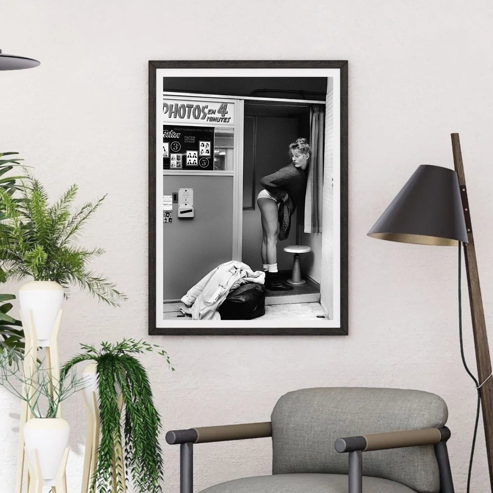 Framed black and white poster of a woman in a Photo Booth on a wall in a modern room with plants and a chair.